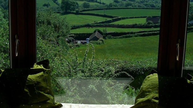 A room with a large window offering a view of rolling green hills and a small house in the distance. - Cosy holiday cottage in the National Park with stunning views and new kitchen. (Widecombe in the moor)