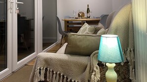 A cozy living room with a sofa, a table with a lamp, a dining table with chairs, and a sliding glass door leading to a balcony. - Cosy holiday cottage in the National Park with stunning views and new kitchen. (Widecombe in the moor)