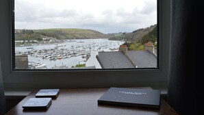 Vista desde una ventana sobre un puerto deportivo con barcos, un paisaje montañoso y un libro titulado “CHARLES HOUSE” sobre un escritorio.