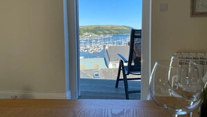 Habitación con vistas a un puerto deportivo y colinas. Hay una silla y una mesa de vidrio con una copa de vino.