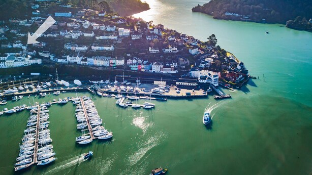 Vista aérea de un pueblo costero con un puerto deportivo, casas y un faro.