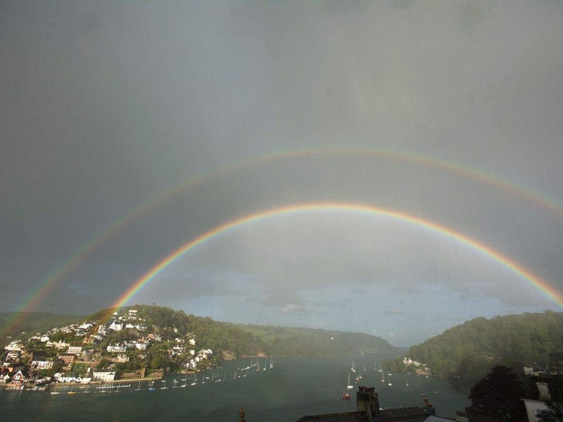 A coastal town with a rainbow arching over the sea, houses along the shore, and boats in the water.
