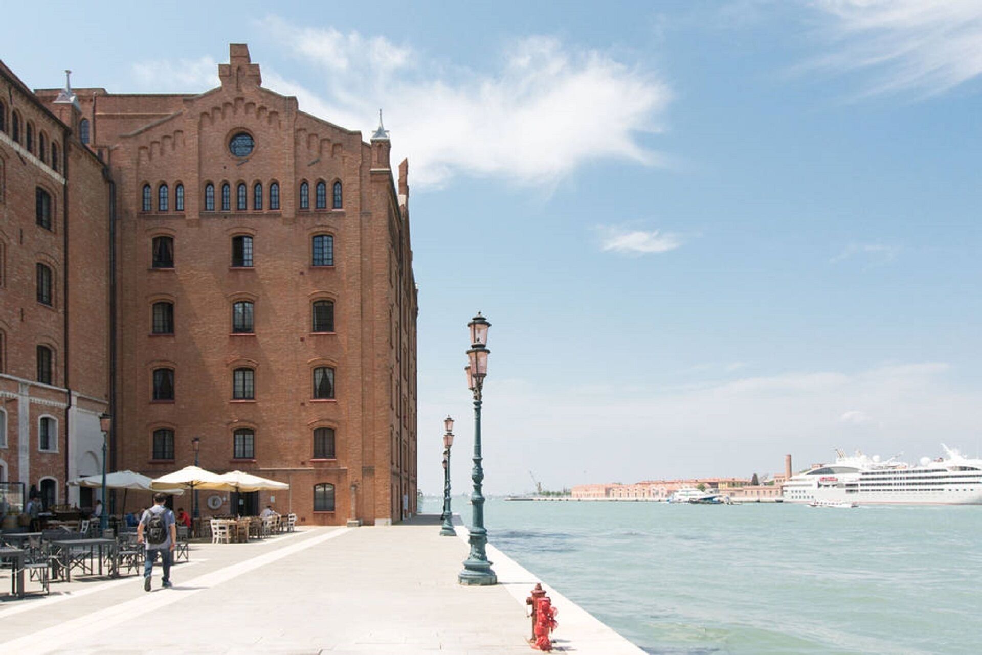 Eine Uferpromenade mit einem historischen Gebäude, Außenplätzen und einem Kreuzfahrtschiff im Hintergrund.