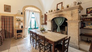Un espace repas comprenant une grande table et des chaises, une cheminée et une vue sur l’extérieur, accessible par un passage en arc de cercle.