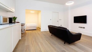 A modern living room with a black sofa, a flat-screen TV mounted on the wall, and a kitchen area with a microwave and a small table with a vase of flowers. - Contemporary in Swiss Cottage, by Veeve (London)