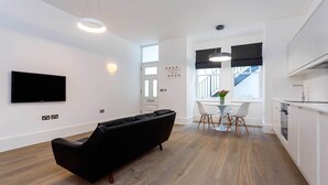 A modern living room with a black sofa, a dining area with a white table and chairs, and a kitchen with white cabinets and a black countertop. - Contemporary in Swiss Cottage, by Veeve (London)