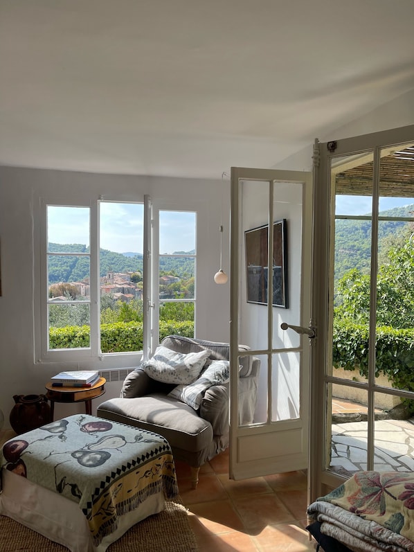 Une pièce dotée d’une grande fenêtre donnant sur un paysage verdoyant et des collines. On y trouve un lit recouvert d’une couverture à motifs, une petite table avec un livre, et une porte qui mène à l’extérieur.