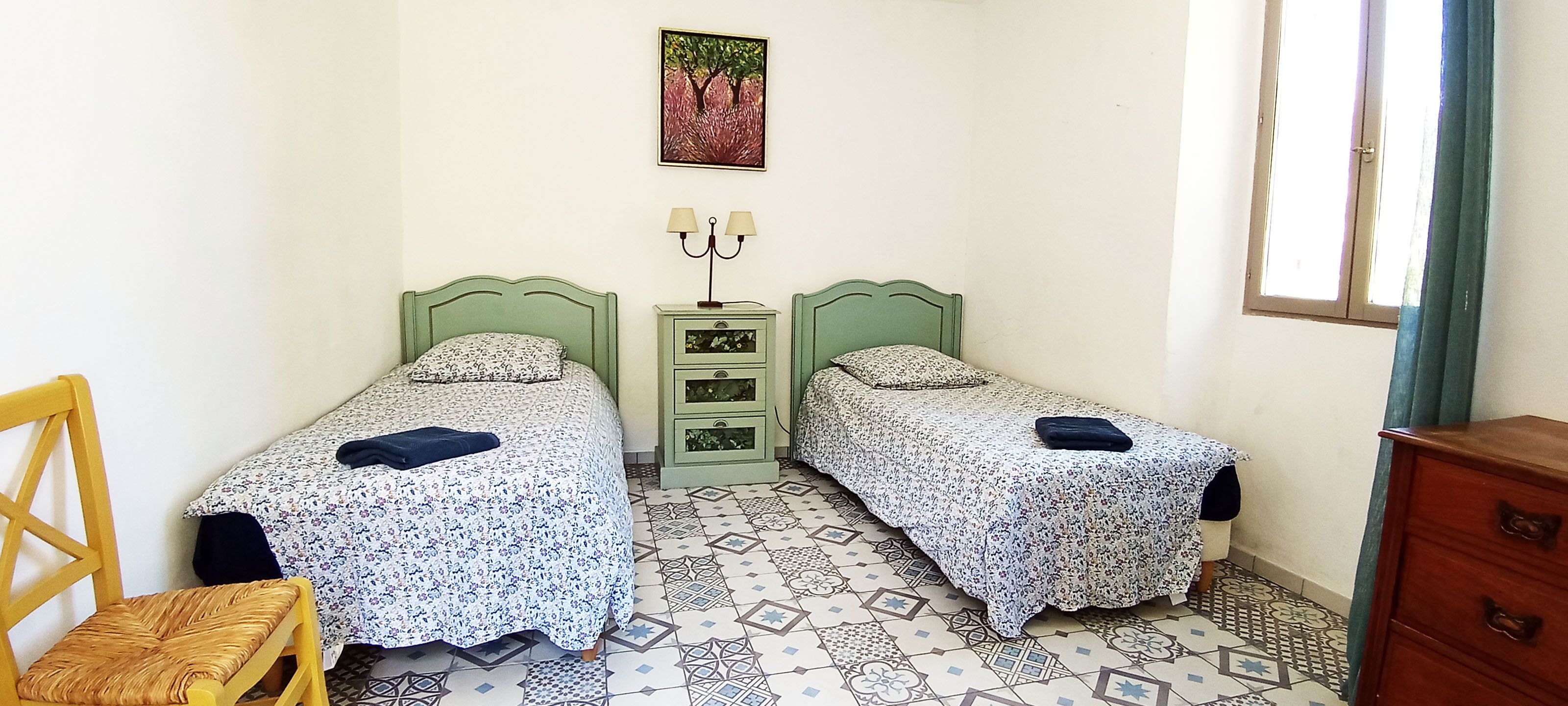 Une petite chambre à coucher avec deux lits simples, une table de chevet avec une lampe, une chaise et une commode.
