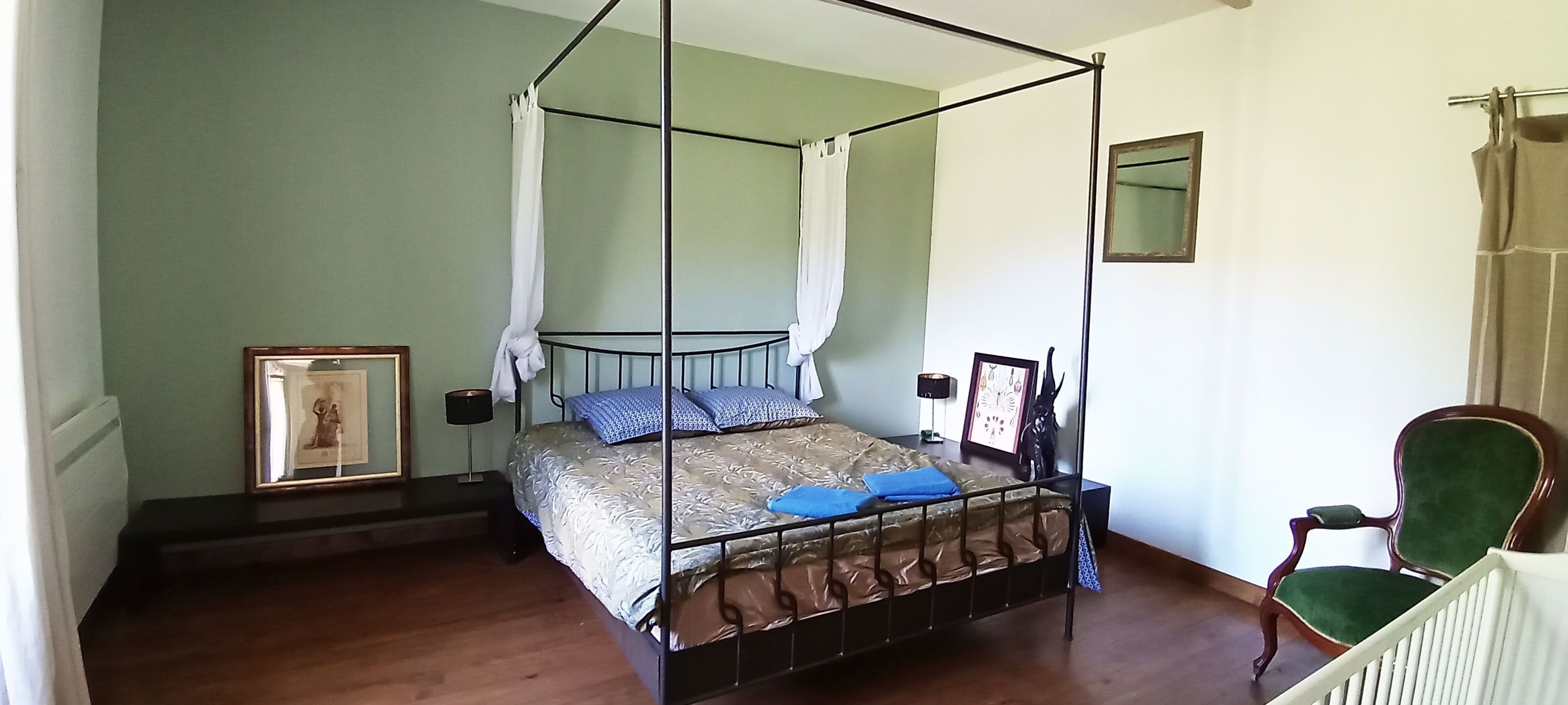 Un lit à baldaquin à quatre poteaux avec une structure en métal, un banc en bois au pied du lit et un fauteuil recouvert d’un tissu vert.
