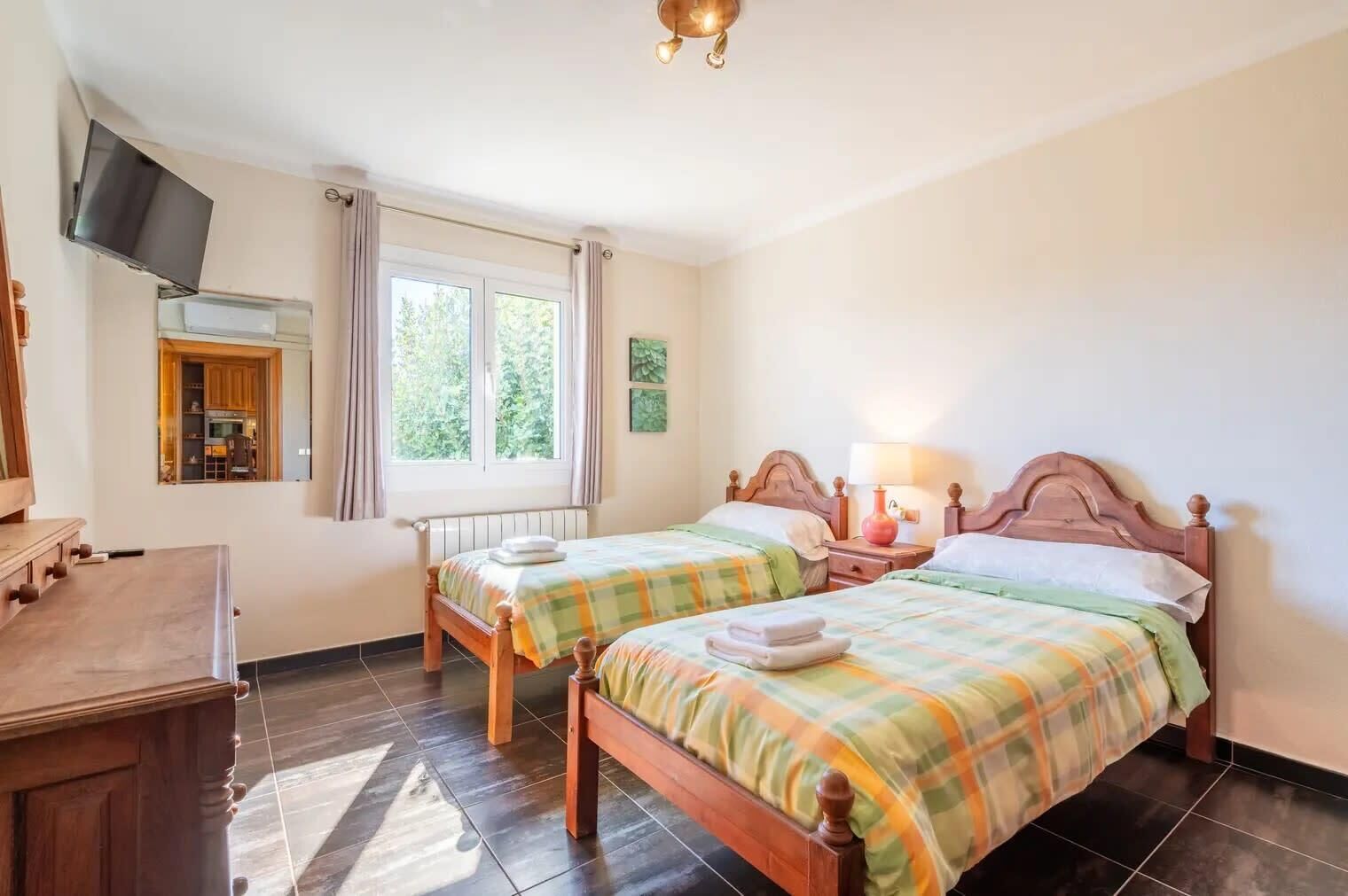 A room with two wooden beds, a TV mounted on the wall, a window with curtains, and a wooden desk.
