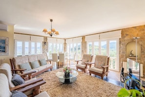 A spacious living room with a large sofa, armchairs, a coffee table, and a chandelier. The room has large windows offering a view of the outdoors. - Hilltop Villa with Panoramic Views over Two Bays (Port d'Alcúdia)
