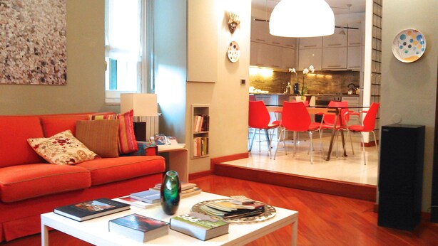 A modern living room with a red sofa, a coffee table with books and a vase, and a view into the kitchen with red chairs.