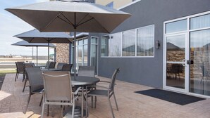 Un patio al aire libre con una mesa, sillas, un paraguas grande y un ventanal de vidrio que da a un espacio interior.