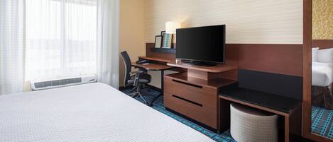 A hotel room with a large bed, a desk with a computer and a flat-screen TV, a chair, a waste basket, and a window with sheer curtains.