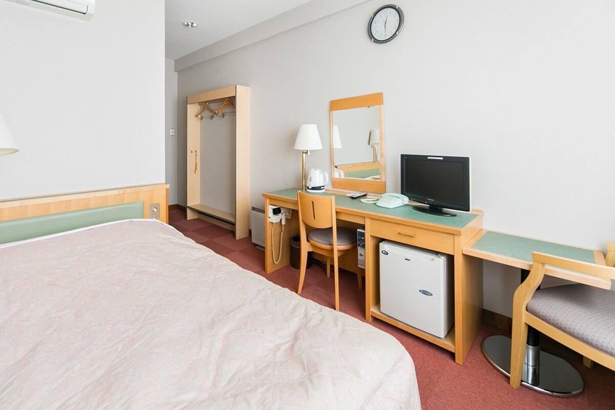 A hotel room with a bed, a desk with a computer, a chair, a wardrobe, and a bedside table with a lamp.