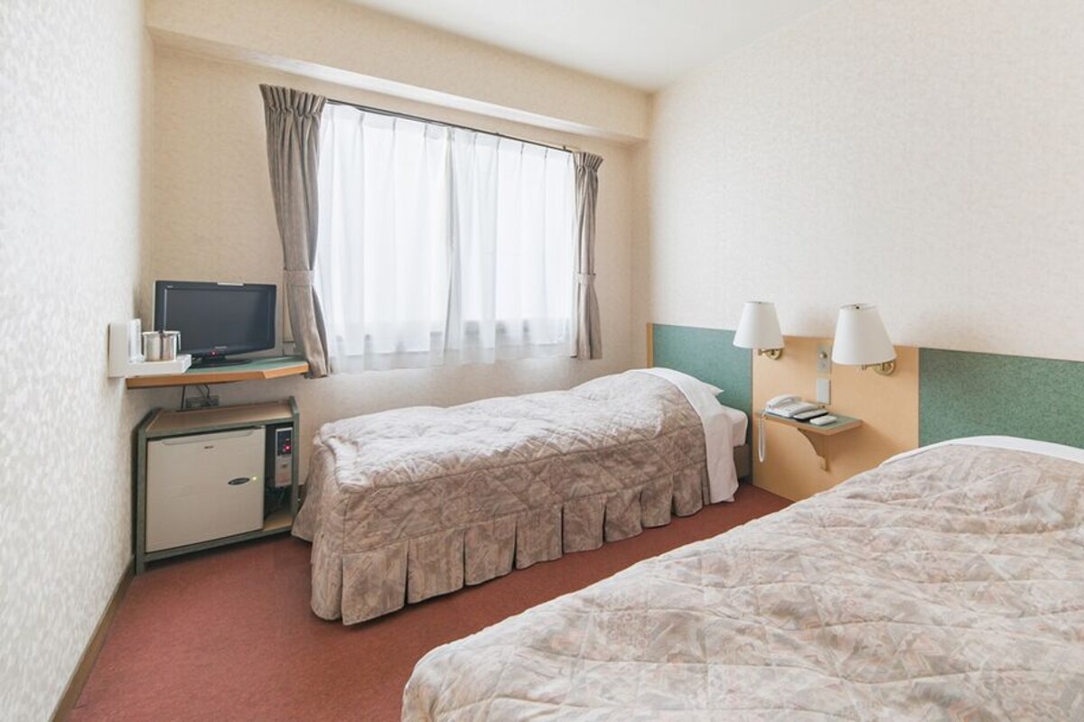 A hotel room with two beds, a desk with a computer, a lamp, and a window with curtains.