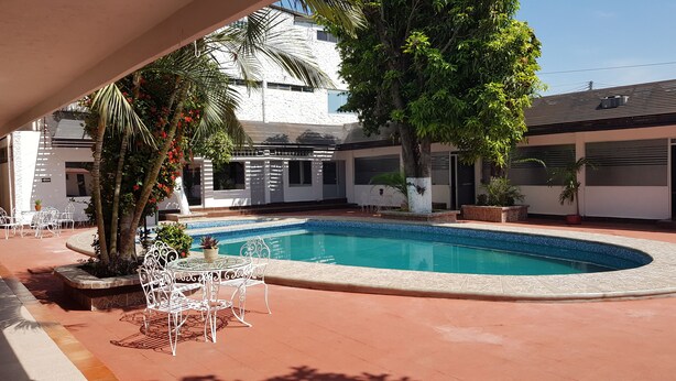 Una piscina con borde de azulejos, rodeada por un patio con sillas y mesas de metal blanco y plantas en macetas.