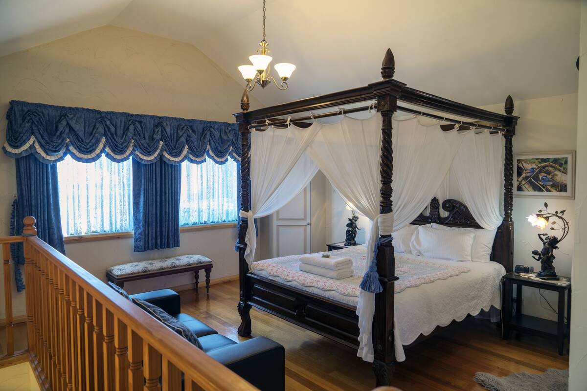 A four-poster canopy bed with white linens, a wooden bench, and a nightstand with a lamp.
