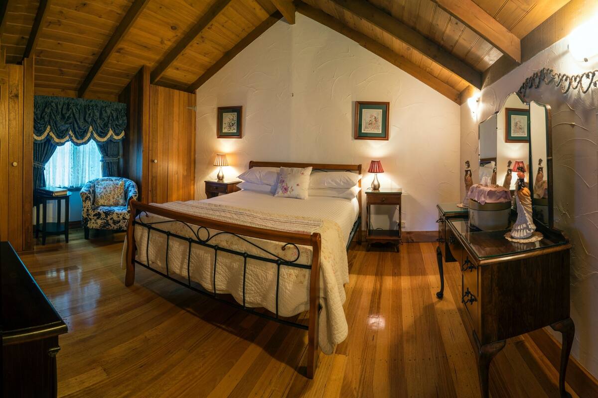 A cozy bedroom with a wooden ceiling, a large bed, two bedside tables with lamps, a chair, and a dresser.