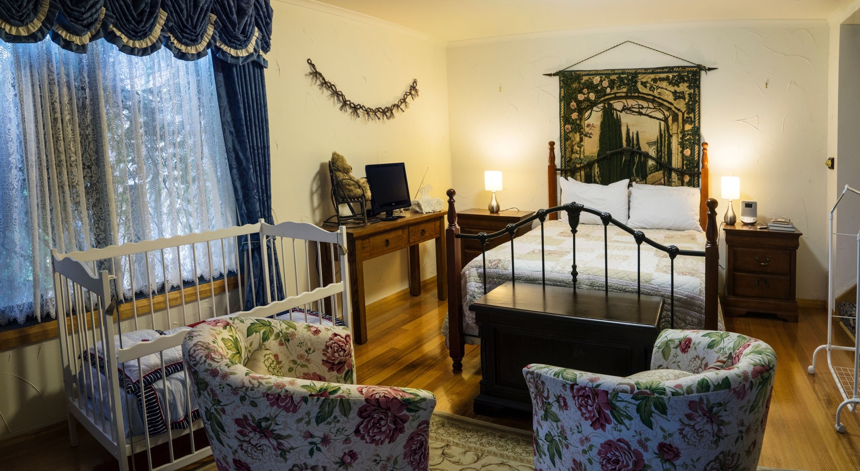 A cozy bedroom with a wooden bed, a crib, floral armchairs, a desk with a computer, and a decorative tapestry.