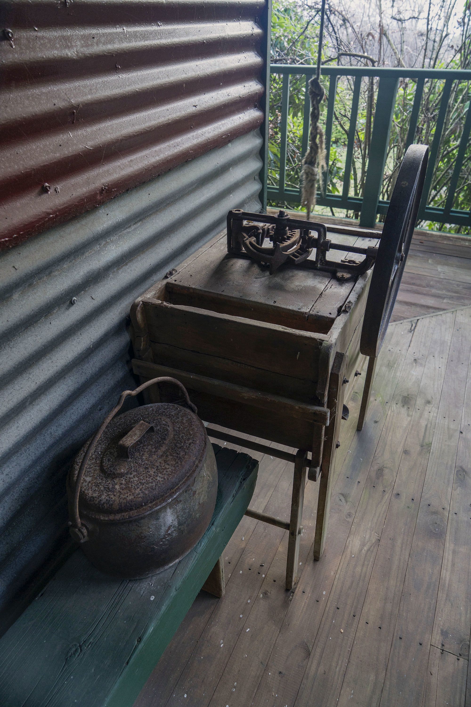 A rustic wooden bench with a metal pot and a mechanical device on it, set against a corrugated metal wall on a wooden deck with a railing.