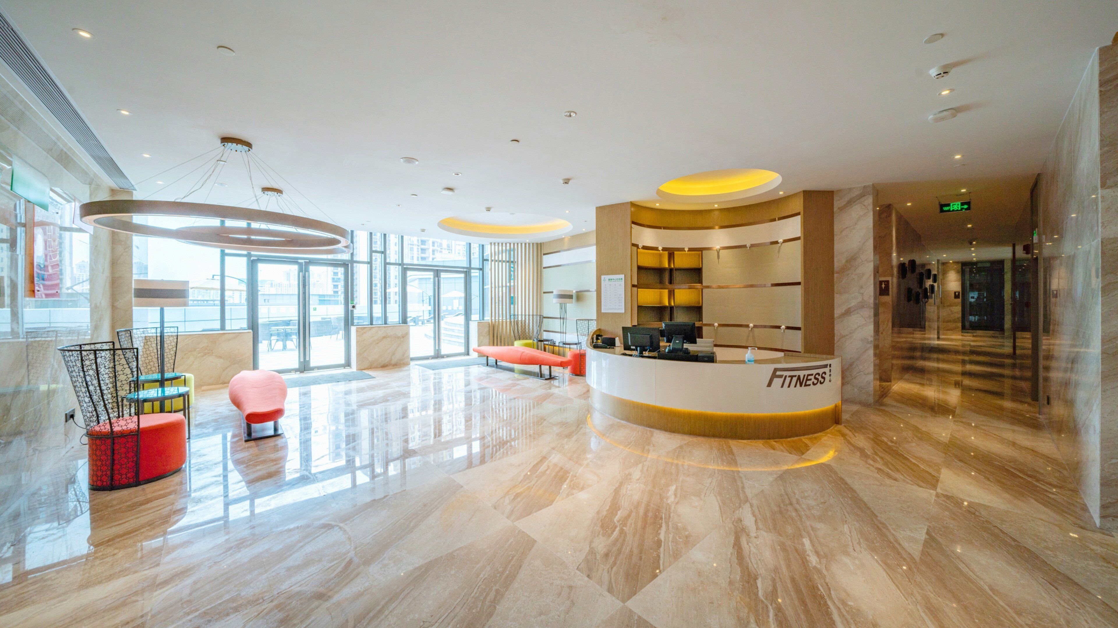 A spacious lobby with a reception desk labeled "FITNESS," modern seating, and large windows offering views of the city.