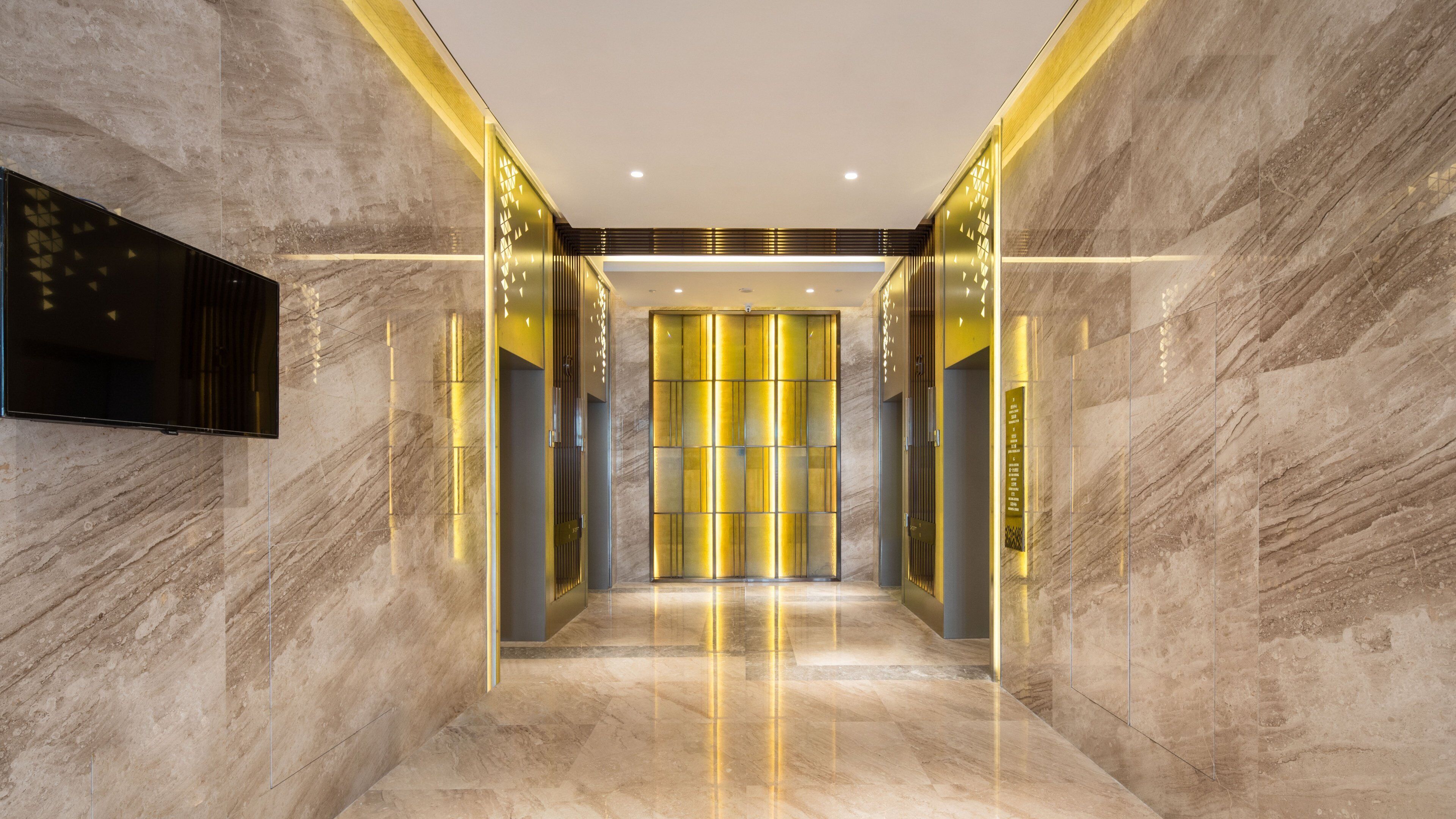 A modern hallway with marble walls, a flat-screen TV mounted on the left, and a bright, illuminated elevator area with a glass door.