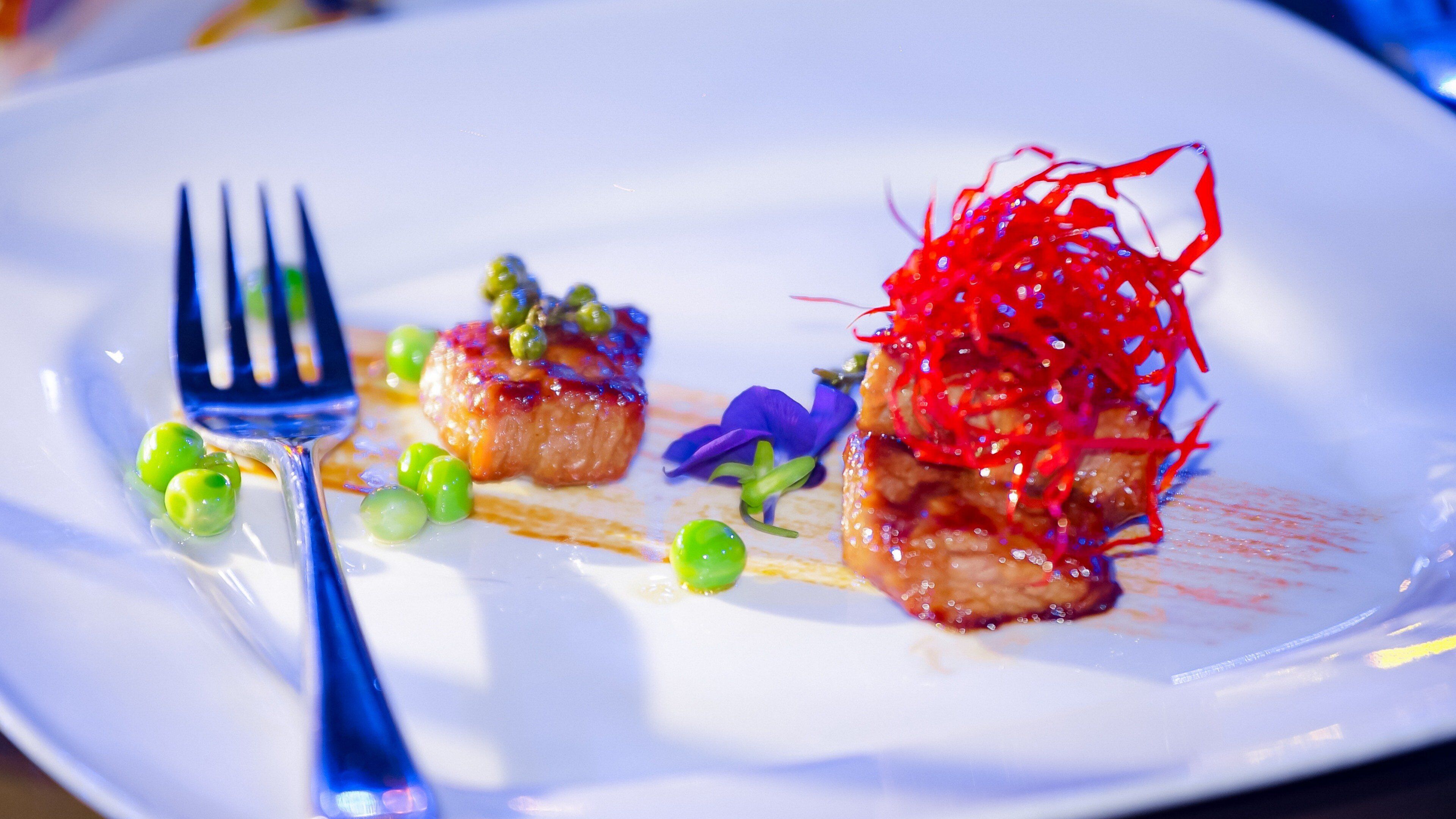Un plato con una variedad de alimentos, incluyendo un trozo de carne, guisantes verdes y una guarnición decorativa.