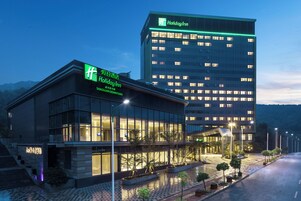 Un hotel Holiday Inn con fachada de vidrio moderna, una entrada bien iluminada y un estacionamiento con árboles.