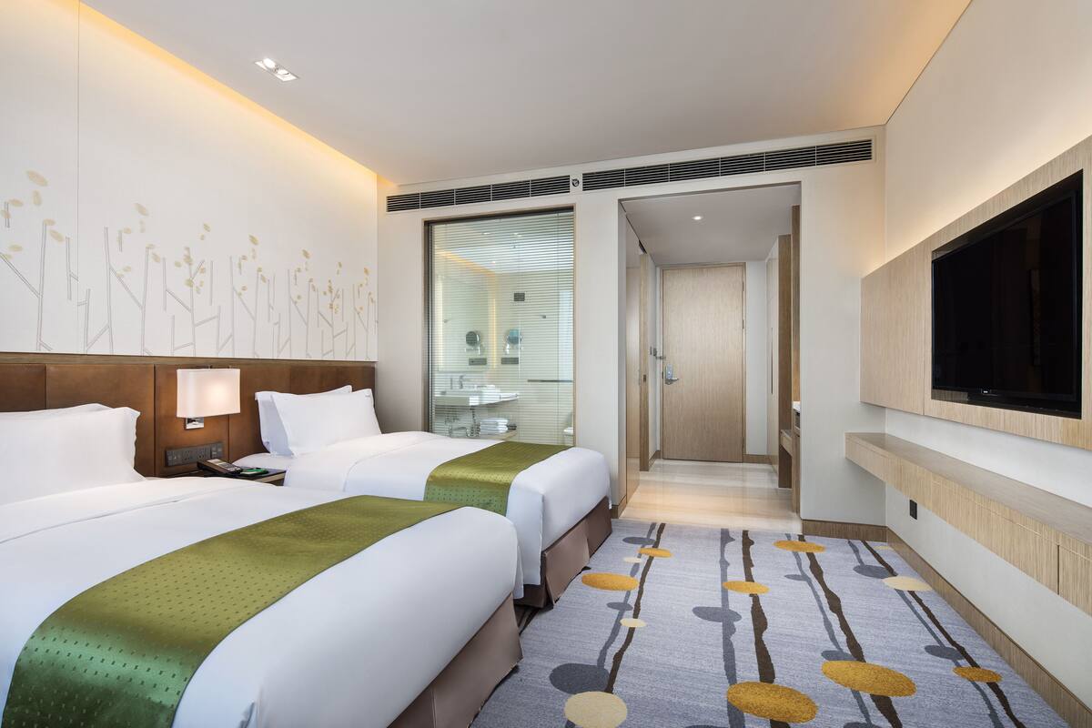 A hotel room with two beds, a TV, a bathroom with a glass shower, and a hallway leading to another room.
