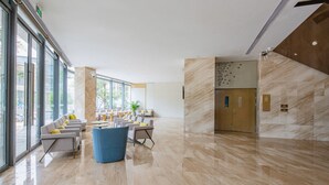 Un hall d’entrée spacieux avec un sol en marbre, un coin salon avec des fauteuils et des tables, de grandes fenêtres et un ascenseur en bois.