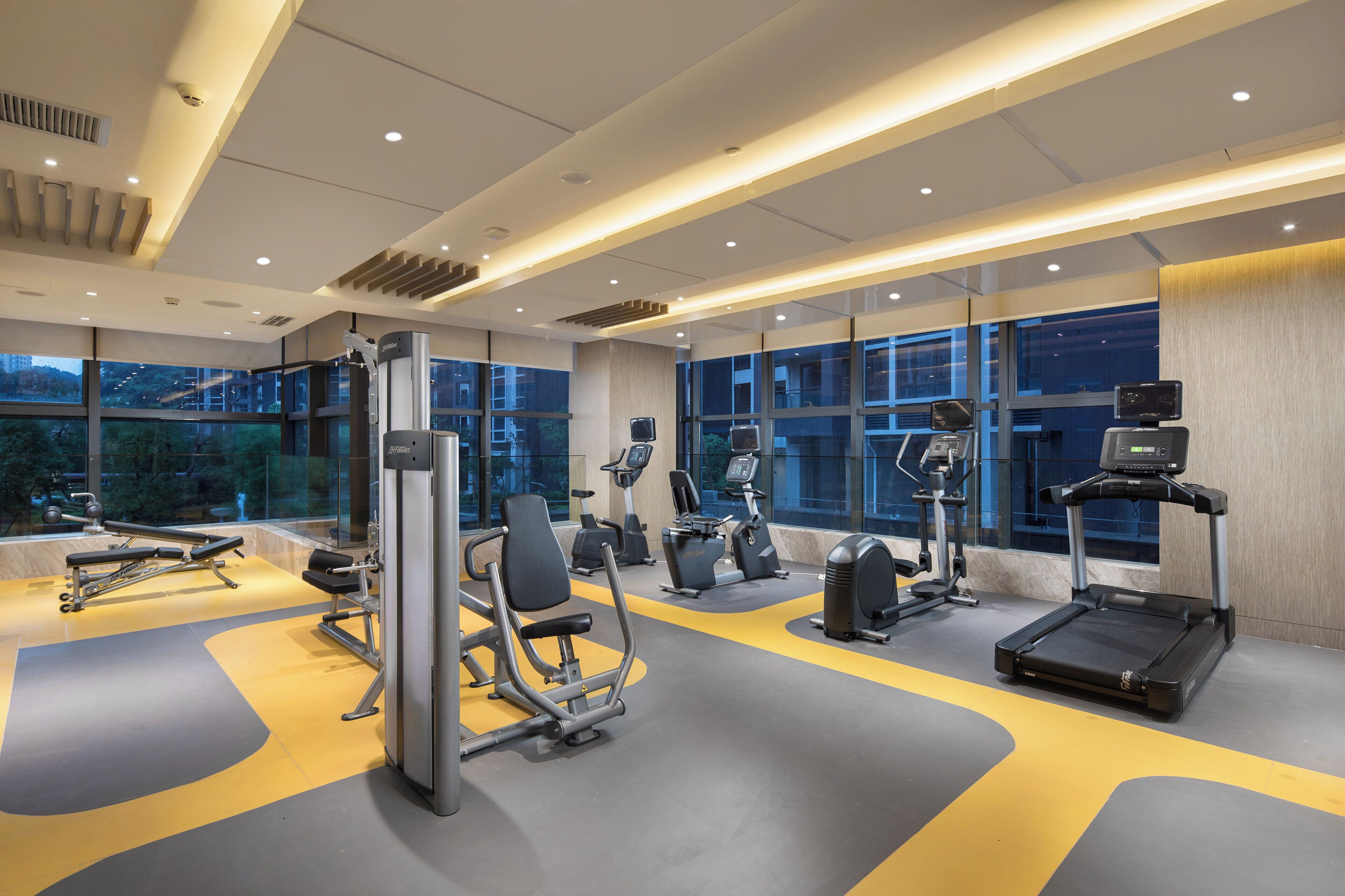 A modern gym with various exercise equipment, large windows, and a yellow and gray floor.