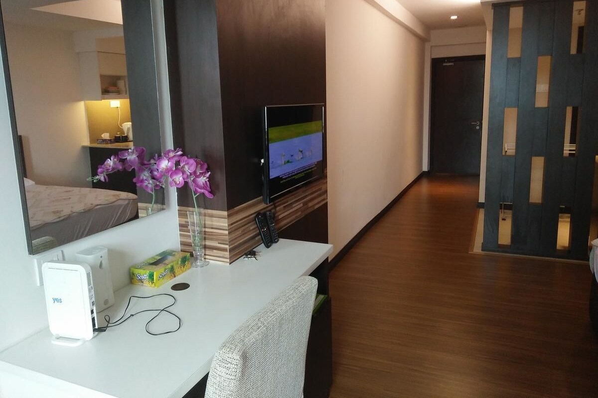 A hotel room with a flat-screen TV, a desk with a computer, a vase with flowers, and a bed in the background.