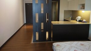 A modern hotel room with a wooden floor, a bed with a floral bedspread, a kitchenette with a sink and a coffee maker, and a dark-colored partition with cutouts. - Best Studio Guest House (Kota Bharu)