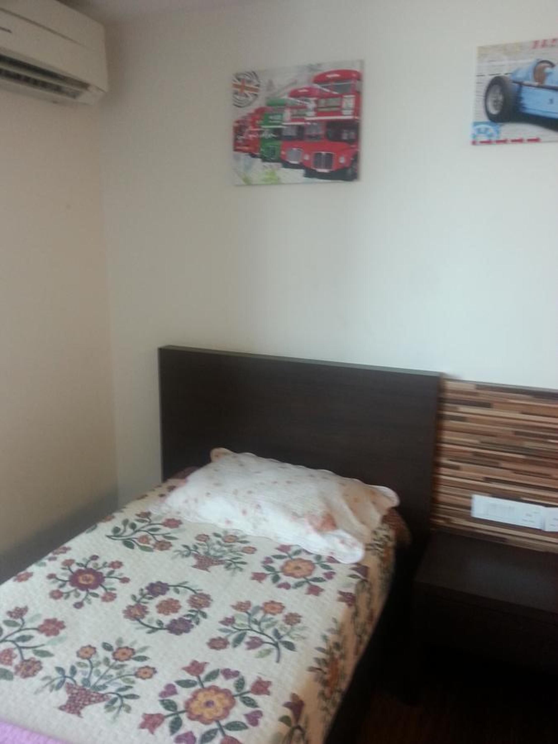 Una habitación pequeña con una cama individual, una colcha de motivos florales, cabecera oscura y un equipo de aire acondicionado de pared.