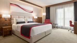 A hotel room with a large bed, two bedside tables, a red armchair, a small round table, and a large window with curtains. - Ramada by Wyndham Beylikduzu (Istanbul)
