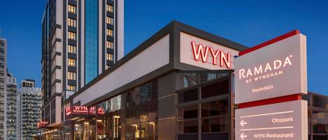 Un hotel moderno con un llamativo letrero de 'RAMADA BY WYNDHAM', un restaurante 'WYIN' y señales direccionales para 'Otopark', 'WYIN Restaurant' y 'Otel Girişi'.