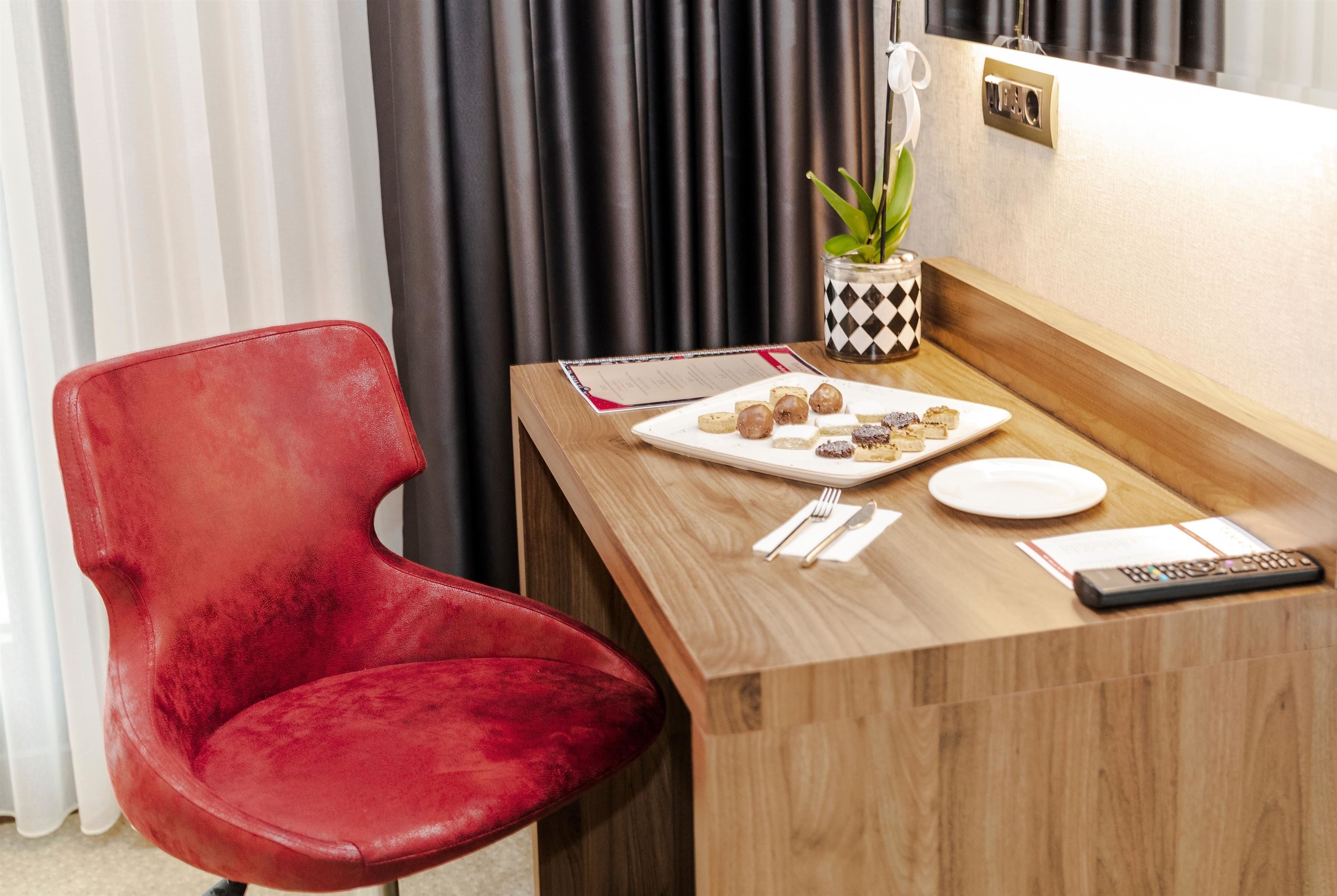 A hotel room with a red chair, a wooden table with a plate of food, a remote control, and a small potted plant.