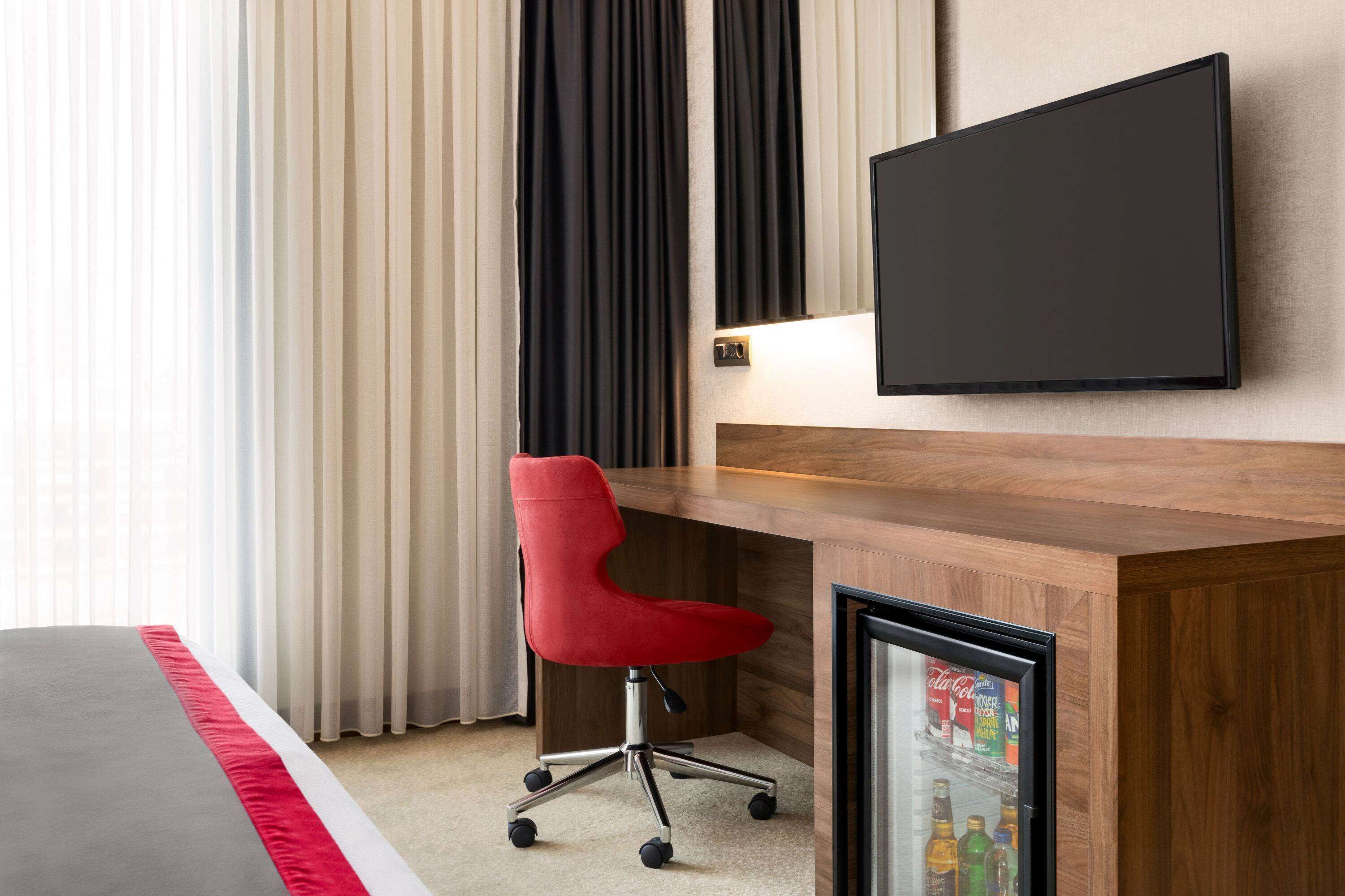Una habitación de hotel con un escritorio de madera, una silla de oficina roja, un televisor de pantalla plana y un minibar.