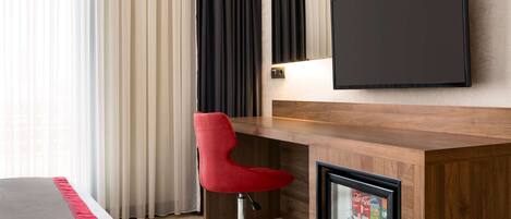 Una habitación de hotel con un escritorio de madera, una silla de oficina roja, un televisor de pantalla plana y un minibar.