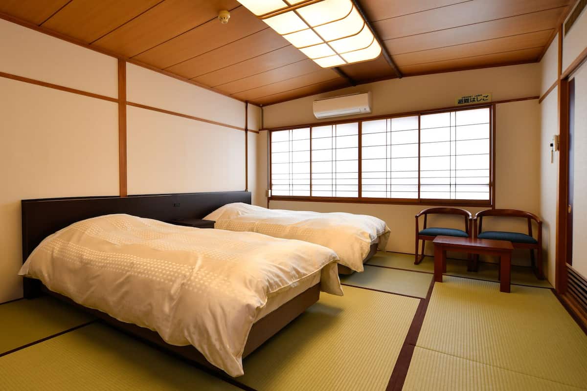 Una habitación japonesa tradicional con suelo de tatami, dos camas con ropa de cama blanca, un escritorio de madera y una ventana con puertas shoji.