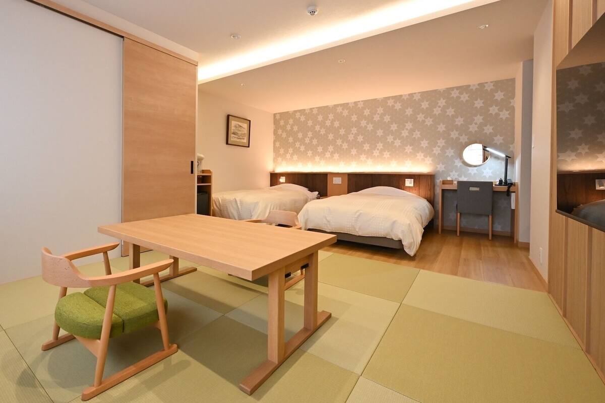 Una habitación de hotel pequeña con un escritorio de madera, dos camas, una mesita de noche y una silla.