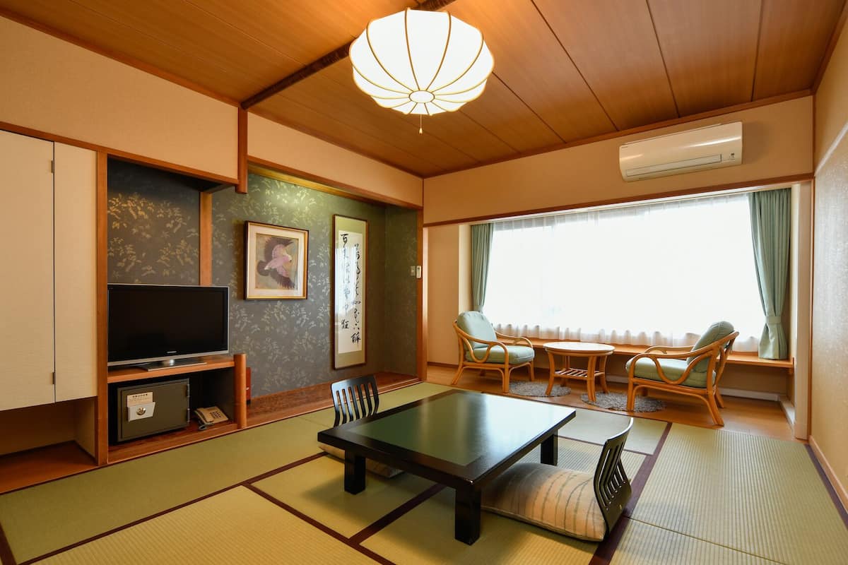 Una habitación tradicional japonesa con suelo de tatami, televisión, estantería, escritorio y ventana con cortinas.