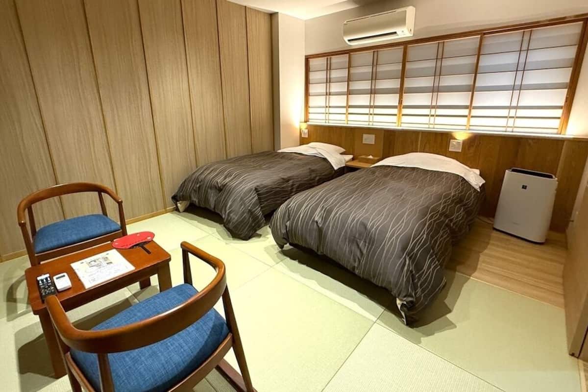 Habitación de hotel con dos camas, una mesita con una silla, aire acondicionado instalado en la pared y un panel corredizo tipo shoji.