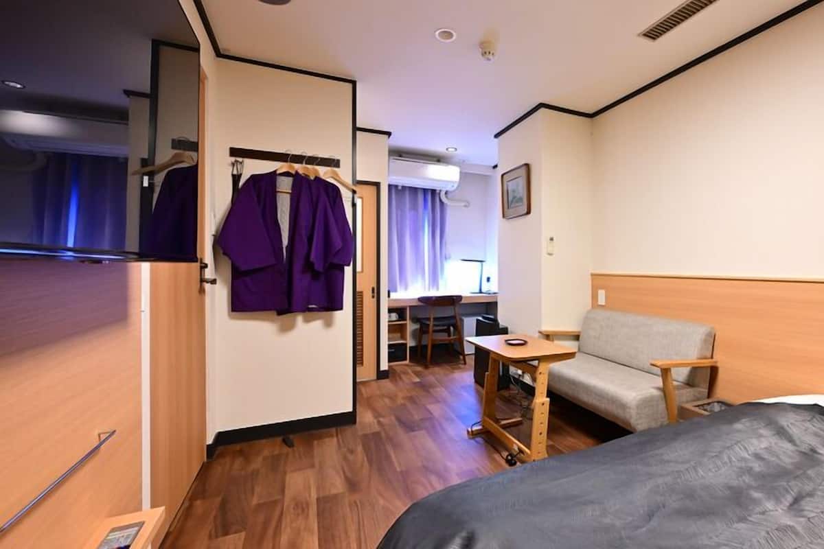 Una habitación de hotel con una cama, un escritorio con una silla, una mesita y un armario con ropa colgada.