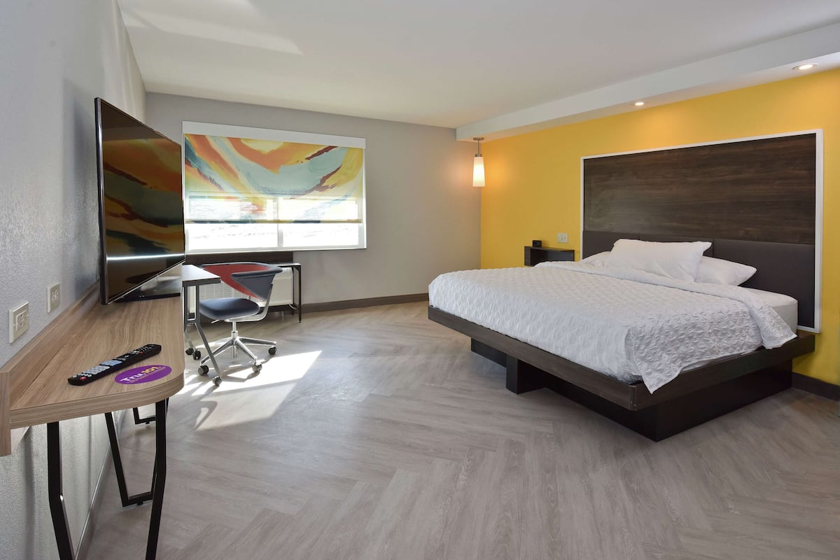 A modern hotel room with a bed, a desk, a TV, and a colorful abstract painting on the wall.