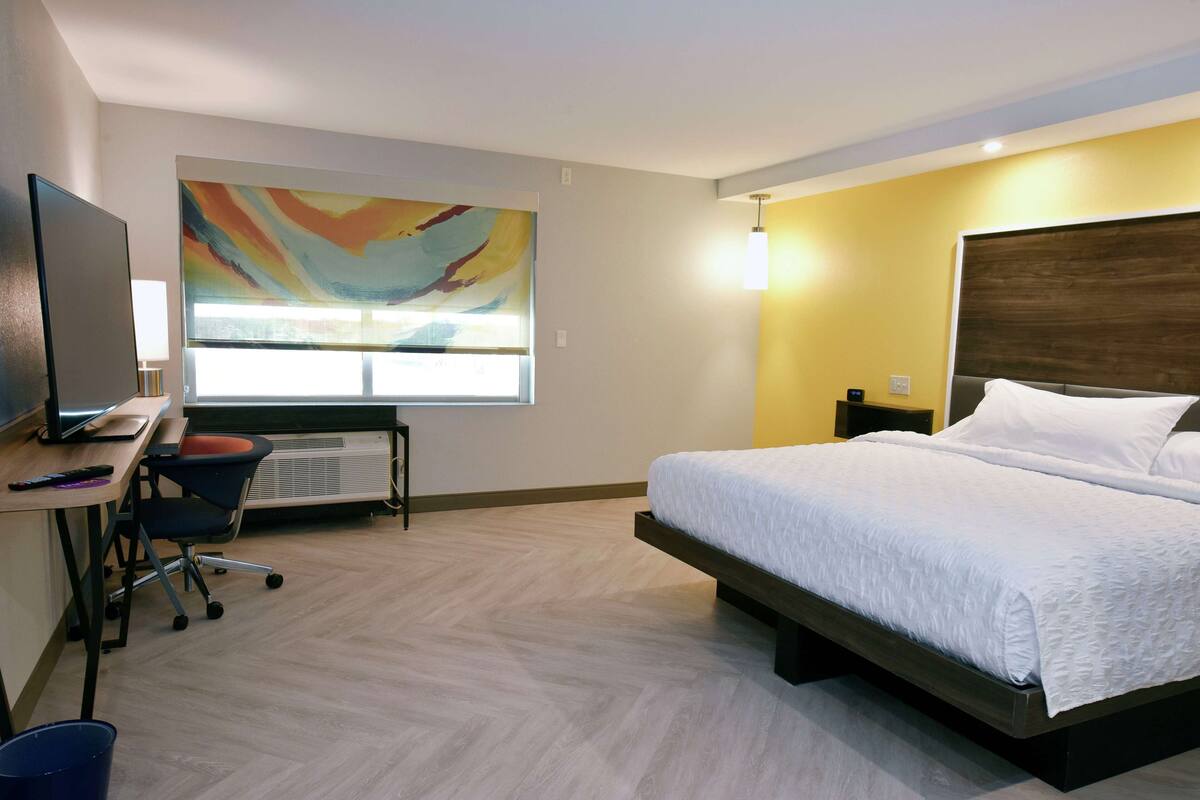 A hotel room with a bed, a desk with a computer, a chair, and a large abstract painting on the wall.