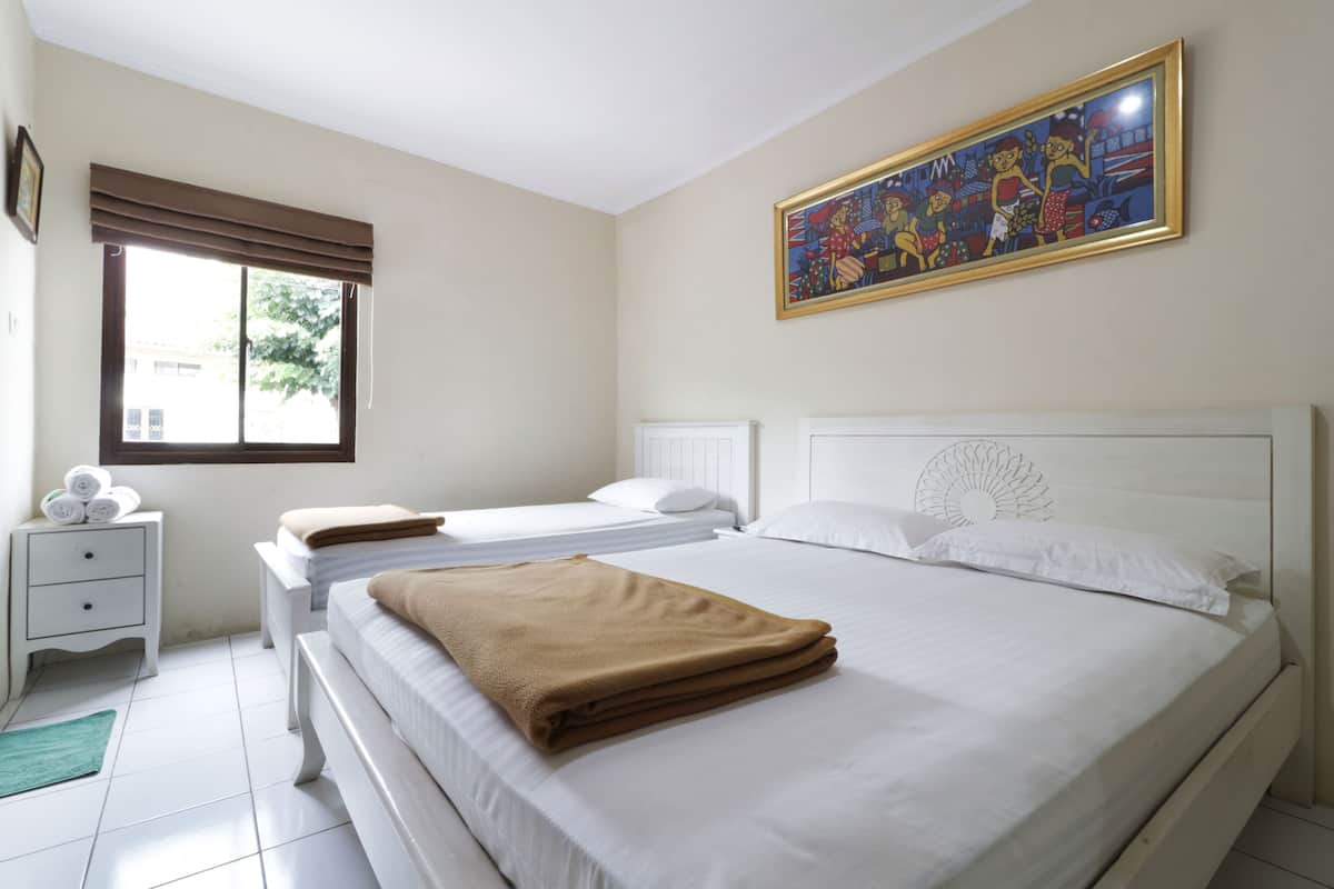 A hotel room with two beds, a window with blinds, a bedside table with a lamp, and a painting on the wall.