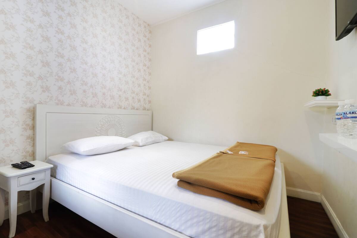 A neatly made bed with white bedding, a small bedside table with a remote control, and a water dispenser on the wall.
