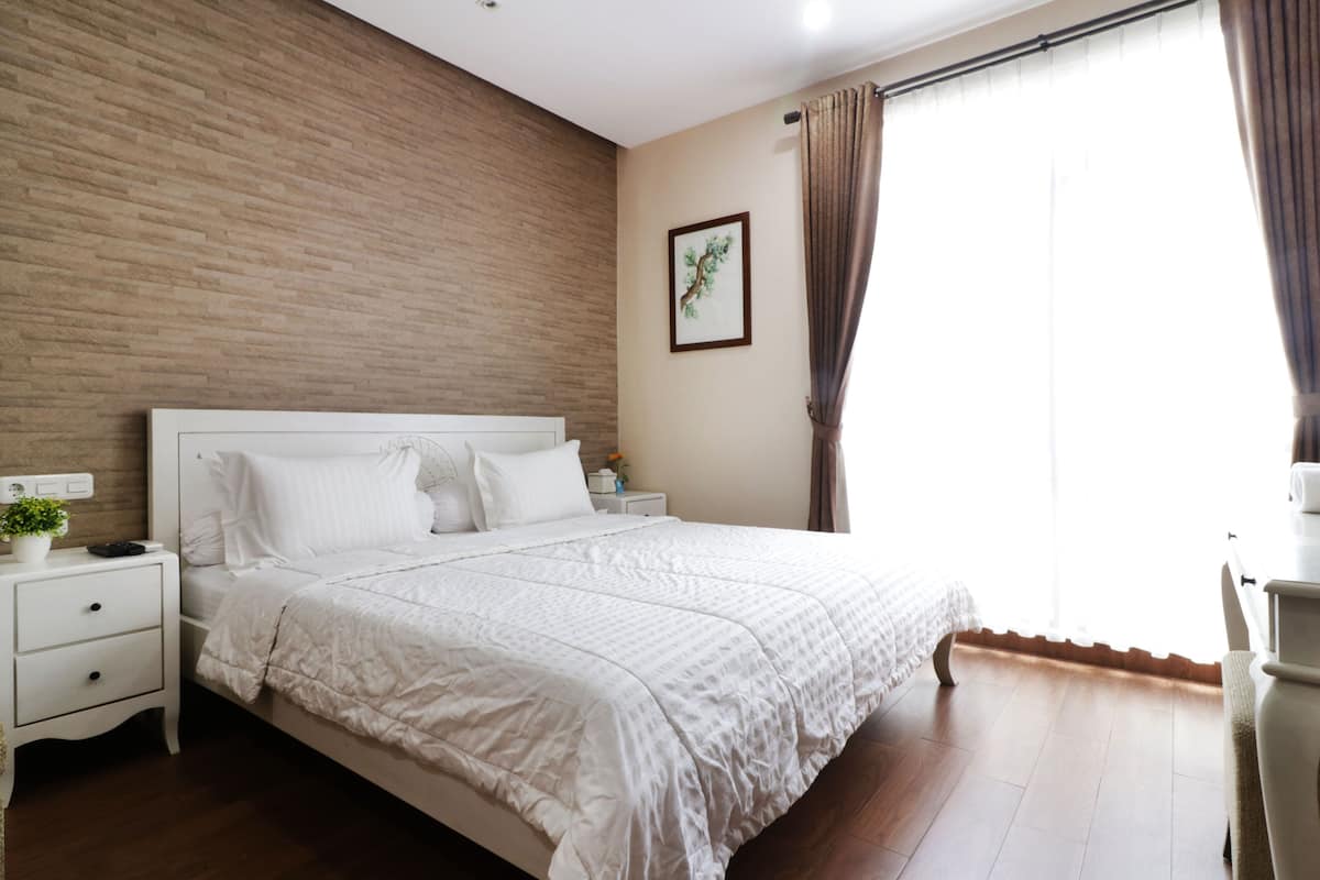 A bedroom with a wooden wall, a bed with white bedding, a nightstand, a small table with a plant, and a window with curtains.